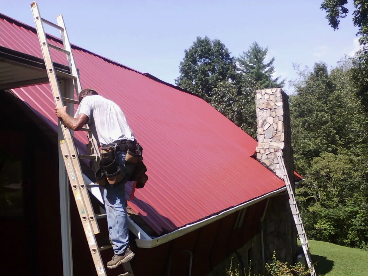 Roofer on a ladder beside a red metal roof and stone chimney — Soffit & Fascia Repair by Roofing Brothers in Conway